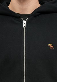 Musternööpidega must kapuutsiga sviiter, millel on känguru tasku, hõbedane tõmblukk ja rindkere piirkonnas väike pruun pitsiga kuldse sarvega ilves.