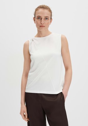 Weißes ärmelloses Oberteil mit einem leichten Drapierungseffekt an einer Schulter, das ein schlichtes Design und eine weiche Textur aufweist. Kombiniert mit schokoladenbraunen Hosen.