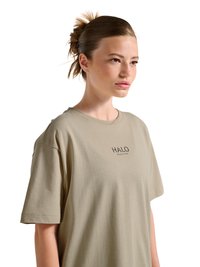 Ung person med samlet hår, iført beige HALO-mærket oversized t-shirt, kigger let mod højre, mod en hvid baggrund.