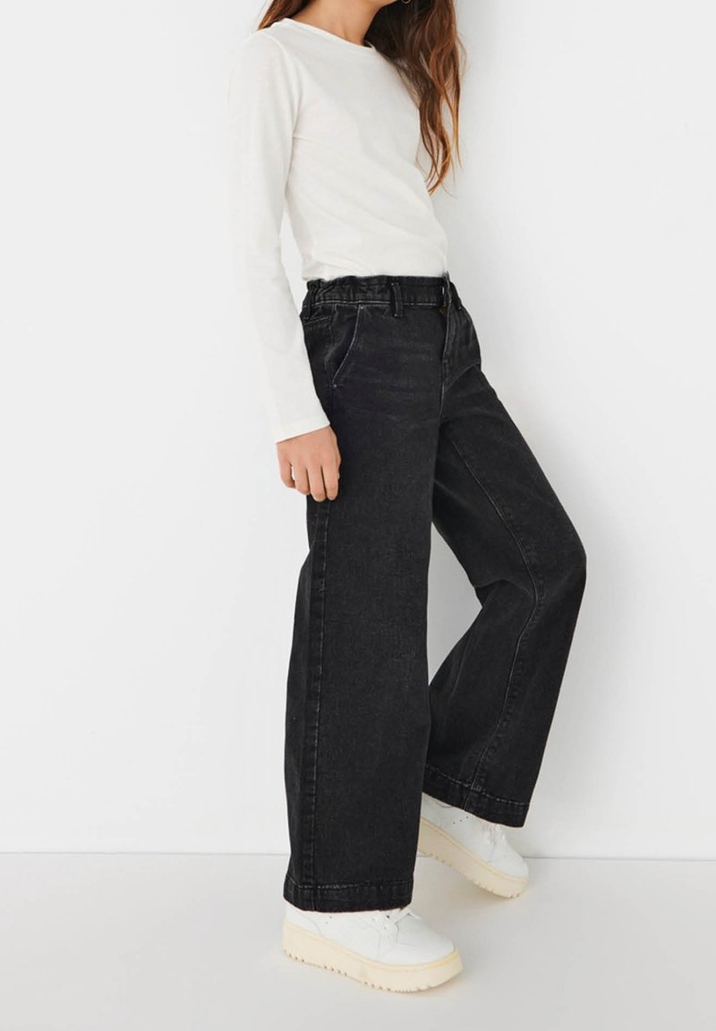 Personne debout portant un haut blanc à manches longues, un jean noir ample taille haute et des baskets compensées blanches sur fond blanc.
