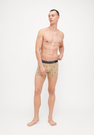 Boxer shorts beige con cinturilla elástica azul marino que presenta patrones de pequeños logotipos repetidos. Tejido suave con un ajuste ceñido, diseñados por encima del tobillo.