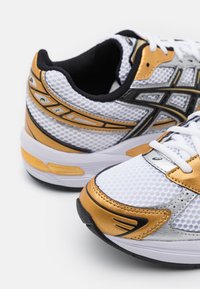 Paire de chaussures de course athlétiques blanches et dorées avec des détails noirs et des lacets blancs sur fond blanc.