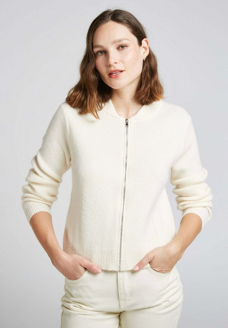 Cardigan zippé crème avec un col rond, des poignets et un ourlet côtelés, ainsi que des poches latérales, fabriqué en tissu doux et texturé.