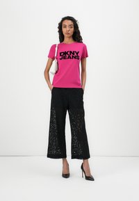 Vaaleanpunainen t-paita, jossa on musta "DKNY JEANS" -teksti, yhdistetty mustiin pitsisiin leveälahkeisiin housuihin ja mustiin korkokenkiin. Asusteina pieni valkoinen laukku.