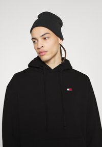 Schwarzer Kapuzenpullover mit einer Fronttasche und einem kleinen, mehrfarbigen Logo auf der Brust. Kombiniert mit einer schwarzen Strickmütze.