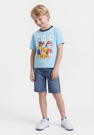 Młody chłopiec ubrany w jasnoniebieską koszulkę z motywem Psiego Patrolu, dżinsowe szorty i białe trampki, stoi i uśmiecha się z lekko pochyloną głową.