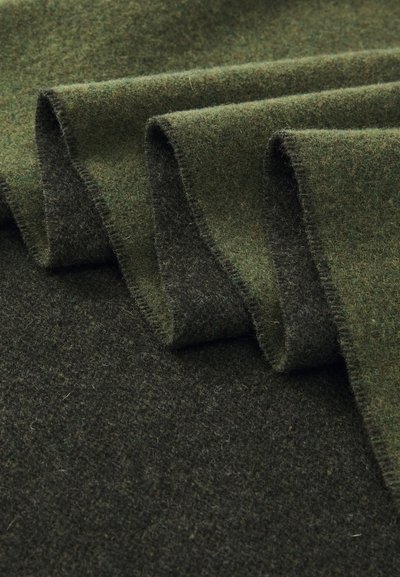 Tissu en laine vert avec une surface douce et texturée, présentant des bords roulés et des nuances de vert variées, créant une apparence superposée.