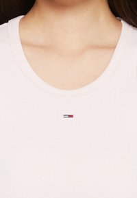 Detailní záběr na klíční kost a krk, oblečený v svetle růžovém ribbovaném triku s kulatým výstřihem, na kterém je uprostřed pod výstřihem malé obdélníkové logo v červené, bílé a námořnické modři.