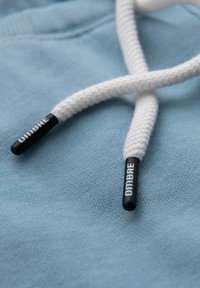 Hvid fletningstræk snor med sorte plastiktipper mærket "DMBRE" hvilende på lyseblåt stof.