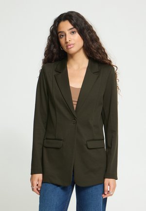 Femme aux longs cheveux bouclés portant un blazer vert foncé, un haut marron et un jean bleu, debout devant un fond clair uni.