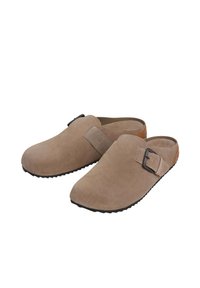Ett par beige mocka slip-on träskor med svarta justerbara spännen och texturerade svarta sulor på en vit bakgrund.