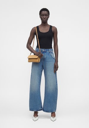 Γυναίκα που φοράει μαύρο τοπ, φαρδιά μπλε τζιν, λευκά μυτερά παπούτσια και κρατάει μια μικρή ταμπά τσάντα χιαστί με ρυθμιζόμενο λουράκι.