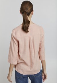 Blouse rose clair à manches longues avec une coupe décontractée, texture en lin doux, encolure ronde et détail de pli au dos, associée à un jean bleu foncé.