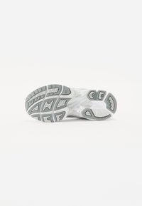 Semelle d'une chaussure de sport blanche avec des motifs de bande de roulement gris et des grips en caoutchouc, présentée sur un fond blanc uni.