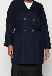 Cappotto trench doppiopetto blu navy con bottoni dorati, cintura e tasche laterali; realizzato in tessuto morbido e testurizzato; vestibilità sartoriale.