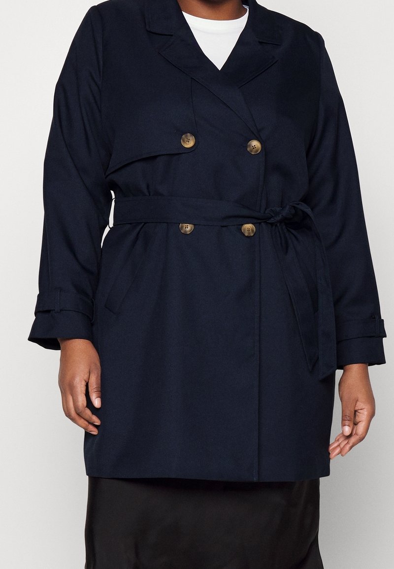 Cappotto trench doppiopetto blu navy con bottoni dorati, cintura e tasche laterali; realizzato in tessuto morbido e testurizzato; vestibilità sartoriale.