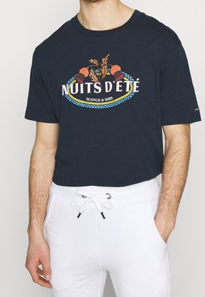 Mężczyzna w czarnym T-shircie z napisem "NUITS D'ÉTÉ SCOTCH & SODA" oraz grafiką żołędzia i liścia, w połączeniu z białymi spodniami ze sznurkiem.