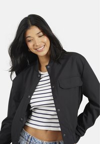 Czarna kurtka zapinana na guziki, z bocznymi kieszeniami i luźnym krojem, noszona na białym i granatowym paskowanym topie typu crop, na jednolitym tle.