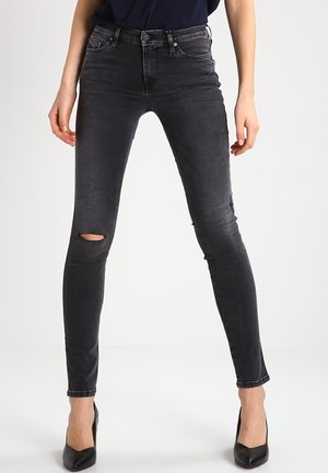 Vrouw draagt zwarte gescheurde skinny jeans en zwarte hoge hakken, staand tegen een effen witte achtergrond.