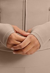 Beige Zip-Up-Oberteil mit gerippten Handgelenkabschlüssen, glatter Textur und figurbetontem Design; die Hände vor der Brust gefaltet, was den Stoff und die Farbe betont.