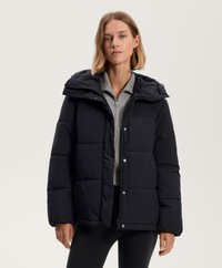 Pufi fekete dzseki kapucnival, patentgombokkal és cipzáras elülső résszel. Szürke felsőn viselve, látható varrásokkal és steppelt mintával.