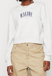 Světle šedá zkrácená mikina s potiskem "Malibu Southern California" v námořnické modři. Obsahuje žebrované manžety a lem, spárované s béžovými kalhotami.