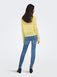 Vrouw met lang golvend haar draagt een gele gebreide trui met getande zoom, blauwe skinny jeans en zwarte enkellaarsjes met hak, met haar rug naar de kijker gekeerd.