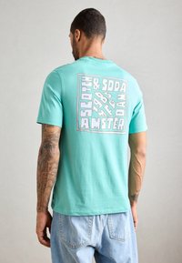 Mand med tatoveringer iført en turkiser t-shirt med hvid grafisk tekst og lyseblå jeans, vist bagfra mod en ensfarvet baggrund.