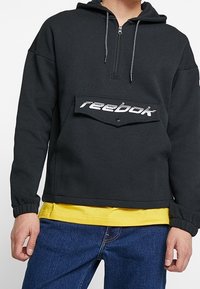 Svart hoodie i mjukt tyg med dragkedja framtill, elastiska ärmslut och en framträdande Reebok-logotyp på bröstet.