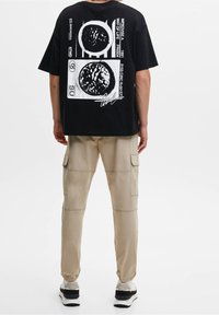 Person, die ein schwarzes Oversize-T-Shirt mit weißen grafischen Druckmotiven und beigen Cargo-Hosen trägt, steht vor einem schlichten weißen Hintergrund.
