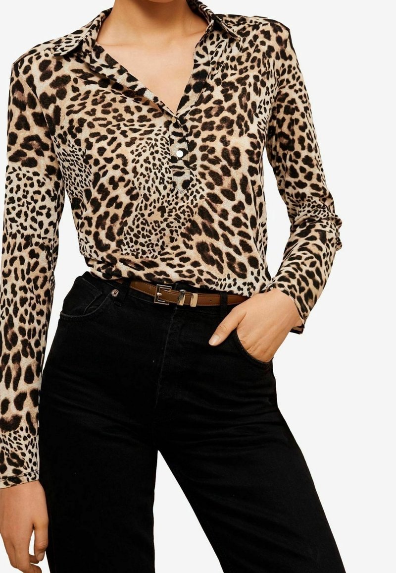 Košile s leopardím vzorem s dlouhým rukávem zasunutá do černých kalhotů s vysokým pasem, s hnědým pásem, jedna ruka v kapse.