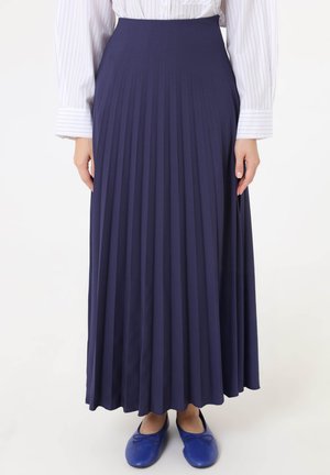 Gonna lunga plissettata blu navy indossata con una camicia bianca a righe e ballerine blu abbinate su una modella in piedi con le mani lungo i fianchi.