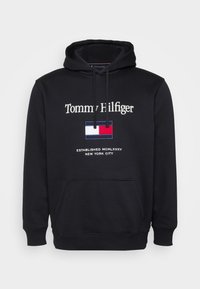 Hanorac negru cu buzunar tip cangur. Prezintă logo-ul "Tommy Hilfiger" și un design cu steagul brodat în roșu, alb și albastru pe partea din față.