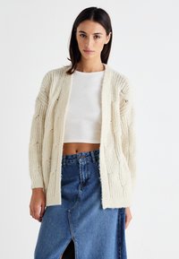 Elfenbeinfarbener Strickcardigan mit Zopfmuster, offener Front und gerippten Manschetten, getragen über einem weißen Oberteil und einem Jeansrock mit Schlitz vorne.