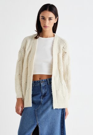 Elfenbensfärgad stickad cardigan med kabelmönster, öppen fram, och ribbade muddar, bärs över en vit topp och en jeanskjol med slits fram.
