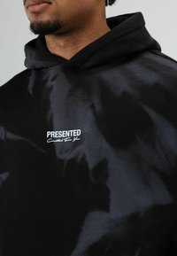 Zwarte hoodie met een glanzende afwerking en een tie-dye effect. "PRESENTED" logo in het wit op de borst, vergezeld door de tekst "Created For You".