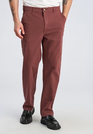 Man draagt roestkleurige rechte broek en zwarte loafers met tassels, handen in de zakken, staand op een effen lichte achtergrond.