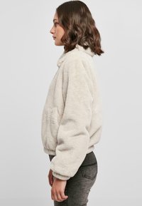 Hvid fleecejakke i blødt plys med høj krave, sidelommer og elastiske manchetter, båret over mørkegrå slim-fit bukser, vist fra siden.