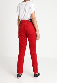 Pantaloni di velluto a coste rossi con vita alta, vestibilità dritta e tasche posteriori visibili. Abbinati a una camicia bianca e scarpe nere.