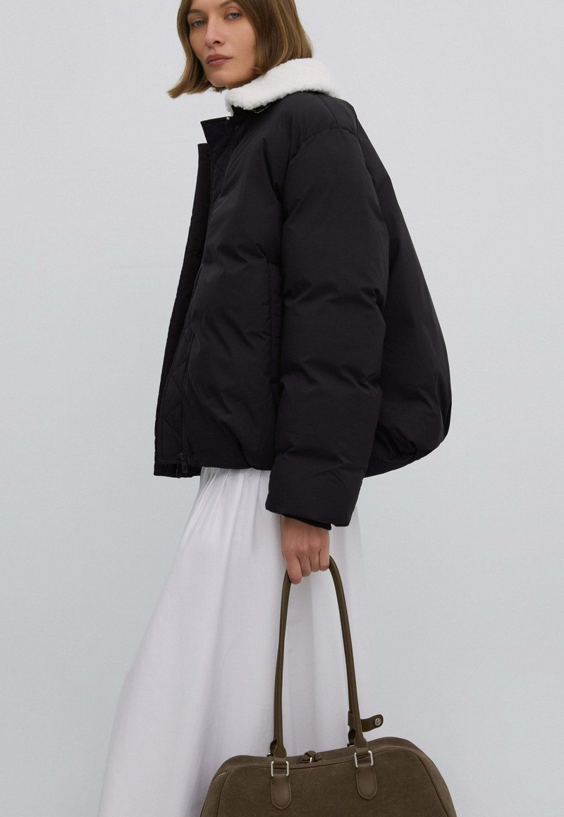 Czarna pikowana kurtka z białym futrzanym kołnierzem, zestawiona z długą białą spódnicą i brązową torebką, wykonana z teksturowanej tkaniny z srebrną galanterią.