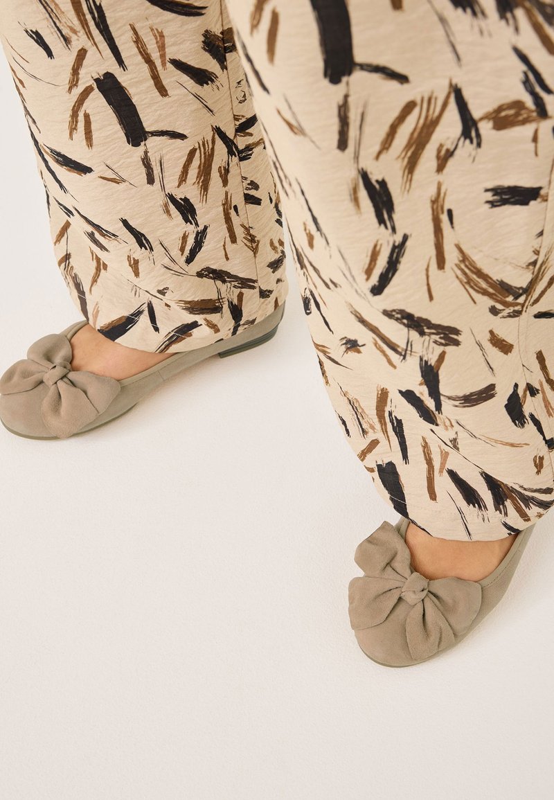 Zapatos planos gris claro con detalle de lazo anudado, combinados con pantalones anchos beige con un patrón abstracto de pinceladas oscuras.