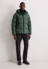 Green puffer jacket with a hood, zip closure, and elastic cuffs. Worn over a black turtleneck and paired with dark pants and black shoes.