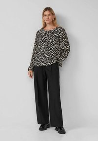 Schwarze Bluse mit langen Ärmeln und weißem gepunktetem Muster, Rundhalsausschnitt, plissierter Front und elastischen Bündchen, kombiniert mit schwarzen, weit geschnittenen Hosen und Schuhen.