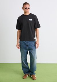 Mand iført sort grafisk t-shirt, løse blå jeans, brune sneakers og solbriller, der står på grøn gulv mod en hvid væg.