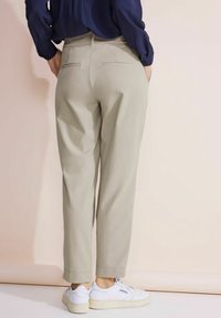 Vaalean beige housut rennoilla mitoilla, joissa on takataskut ja käänteet lahkeissa, yhdistettyinä valkoisiin lenkkareihin. Kankaan pinta on sileä.