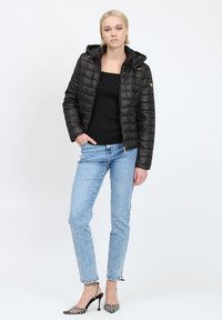 Giacca puffer nera trapuntata con cappuccio, chiusura frontale con zip e dettagli dorati, indossata sopra un top nero, abbinata a jeans azzurri chiari e tacchi con fantasia.