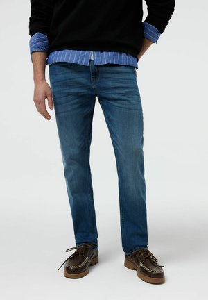 Mężczyzna w niebieskich obcisłych dżinsach, brązowych skórzanych butach, niebieskiej koszuli w paski i czarnym swetrze z podwiniętymi rękawami.