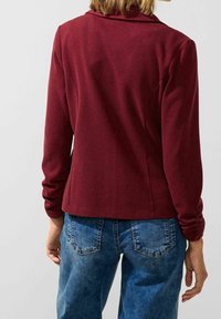 Burgundská bunda s dlouhým rukávem a měkkou texturou, s širokým límcem a přiléhavým střihem, v kombinaci s modrými džínami.