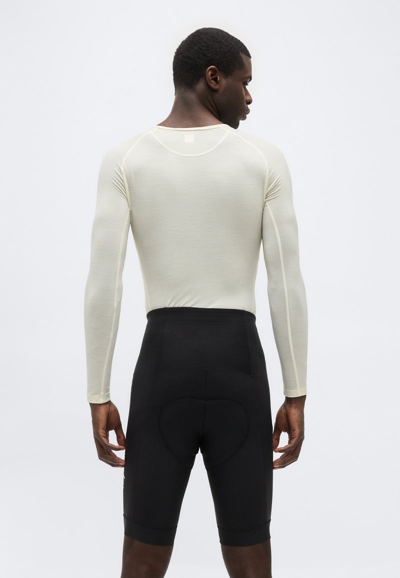 Man wearing a fitted long-sleeve beige top and black padded cycling shorts, standing facing away against a plain gray background.