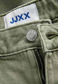 Κοντινό πλάνο φωτεινών πράσινων τζιν denim με ετικέτα JJXX, μεταλλικό κουμπί χαραγμένο με JJXX και μερικώς ανοιχτό φερμουάρ.